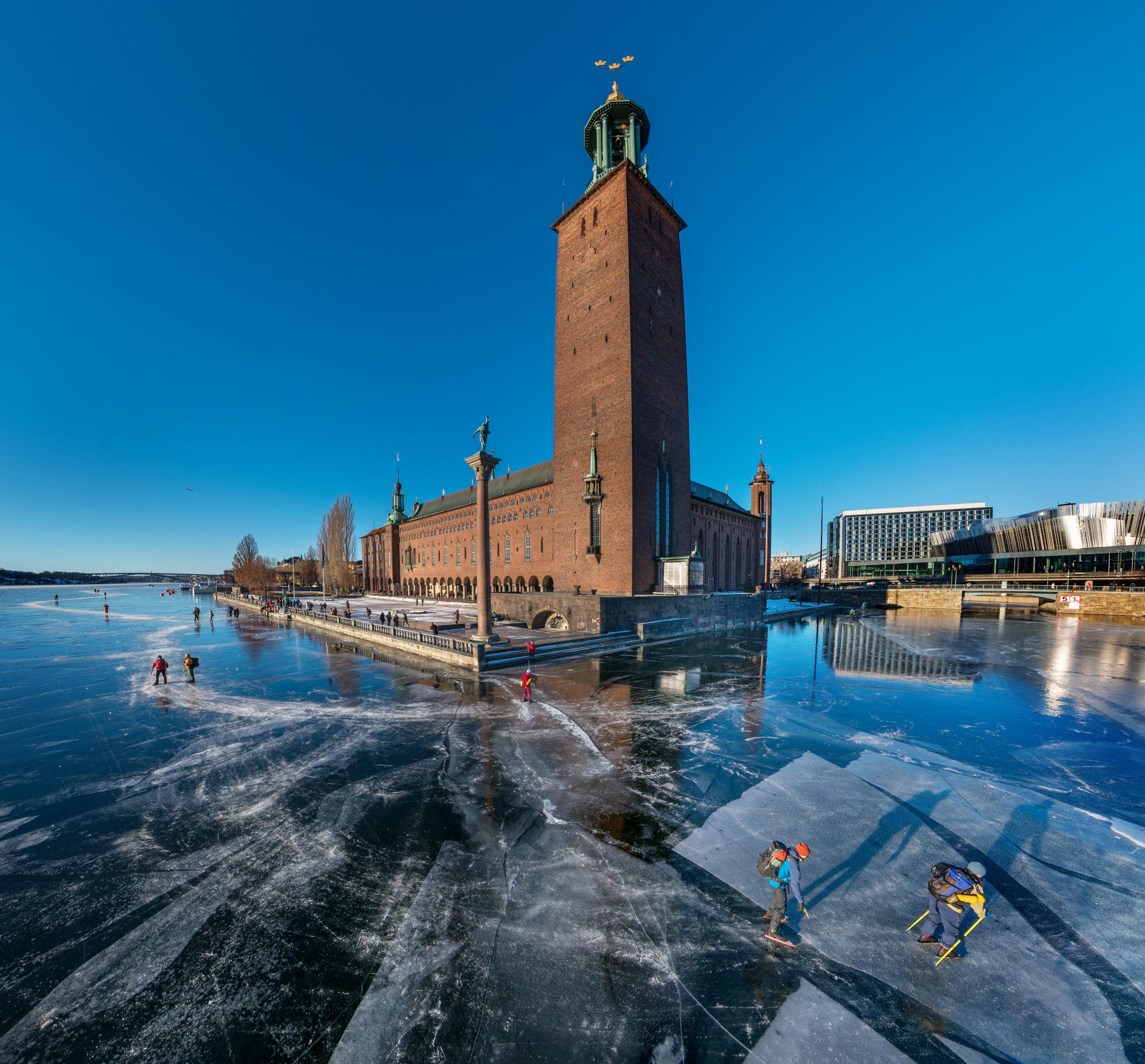 People ice skate around a large brick building with a huge clock tower.