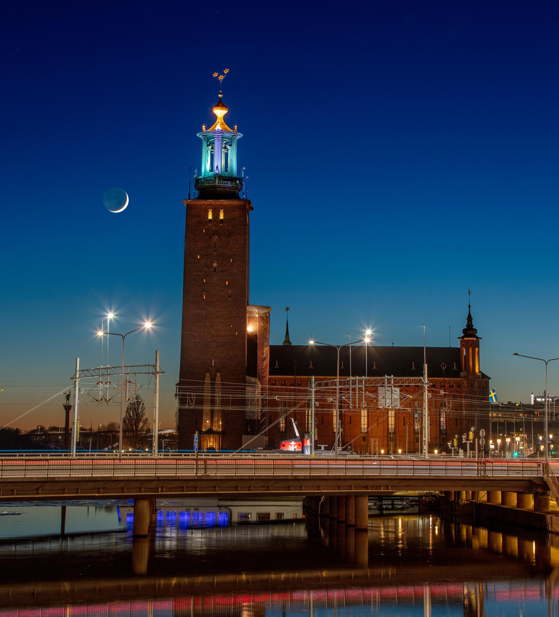 The new moon shines behind a large brick building with a huge clock tower.