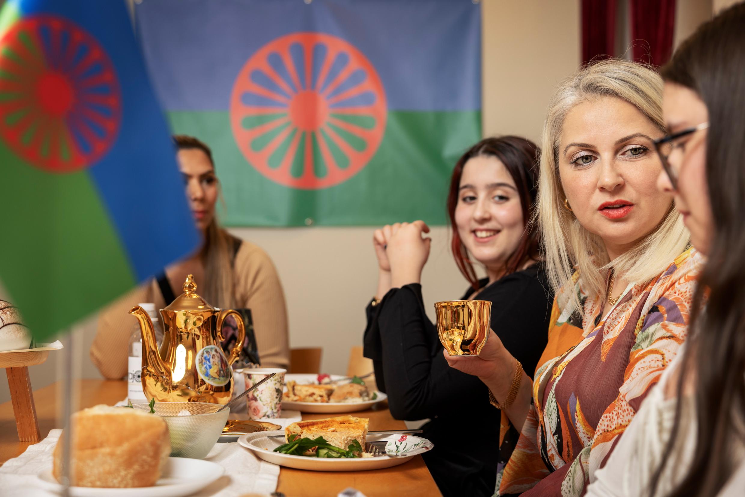 Four women sit around a table eating pie and drinking tea. Roman flags are visible in the picture.
